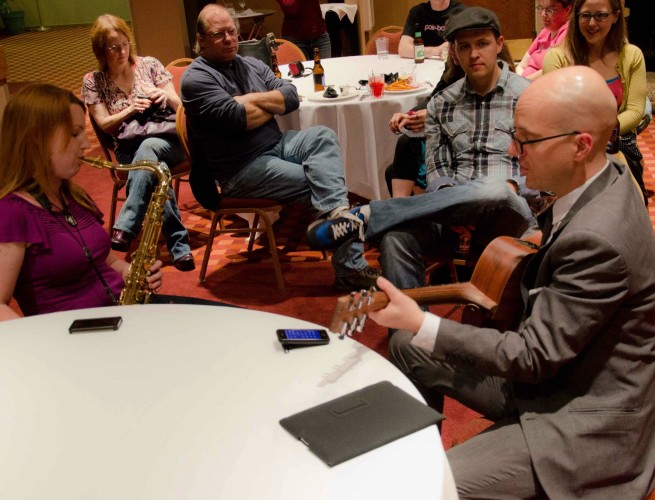 Ashley plays saxophone and George plays guitar near a table for a small seated audience