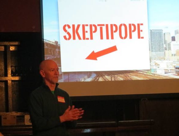 Jairus standing next to a screen that has an arrow pointing to him with the caption "SKEPTIPOPE"